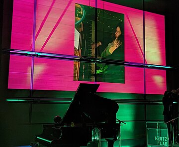 Dunkle Bühne mit zwei Flügeln und einer Pianistin, die über eine Leinwand zu sehen ist