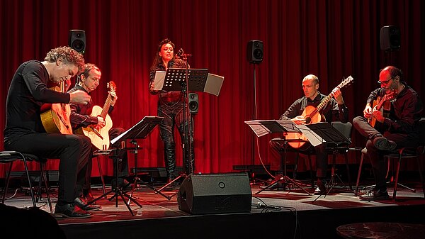 Vier Gitarrenspieler mit Sängerin auf einer Bühne vor einem roten Vorhang