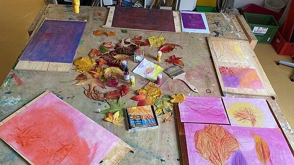 Blick von oben auf einen Holztisch, auf dem bemallte Papiere, Herbstblätter, Pinsel und Farben stehen.