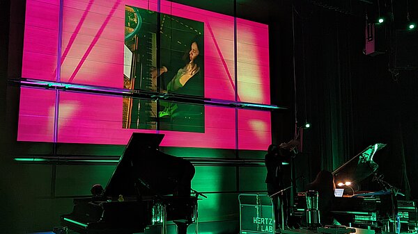 Dunkle Bühne mit zwei Flügeln und einer Pianistin, die über eine Leinwand zu sehen ist